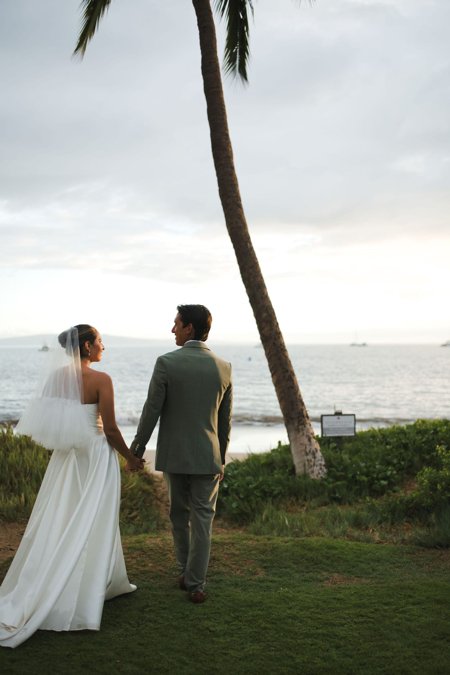 Bride and groom holding hands walking toward ocean at tropical beach wedding with palm tree at sunset.