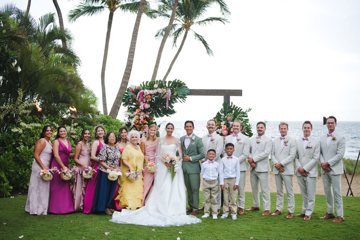 Wedding party poses outdoors in Hawaii with palm trees, ocean backdrop, bridesmaids in colorful gowns and groomsmen in tan suits.