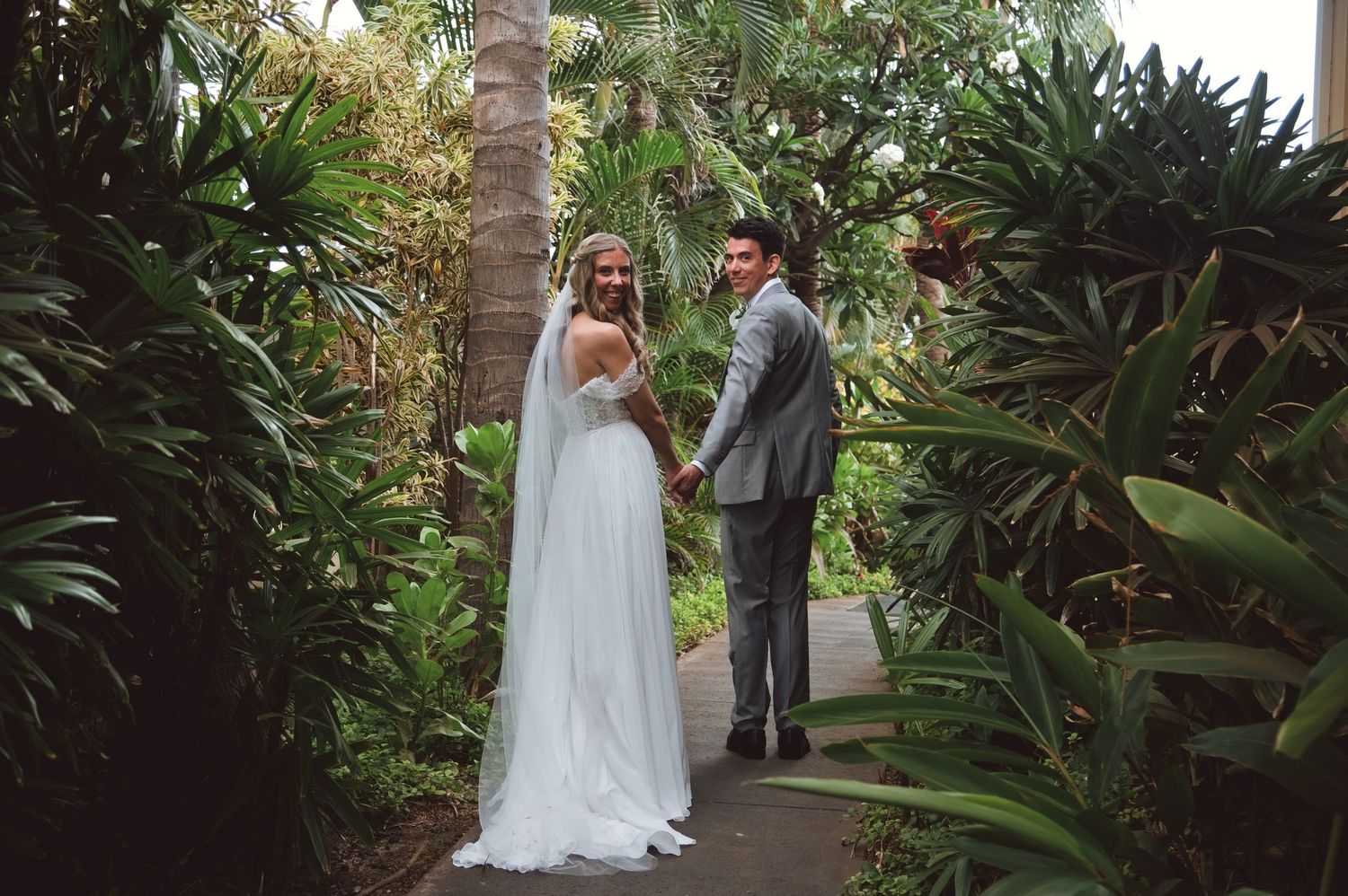 Bride and groom holding hands on a tropical garden path, surrounded by lush greenery and palm trees.
