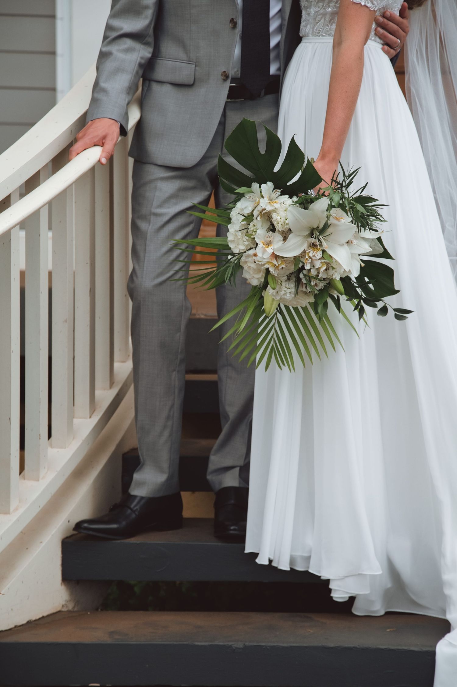Bride in white gown holding tropical bouquet with white flowers stands beside groom in gray suit on staircase.