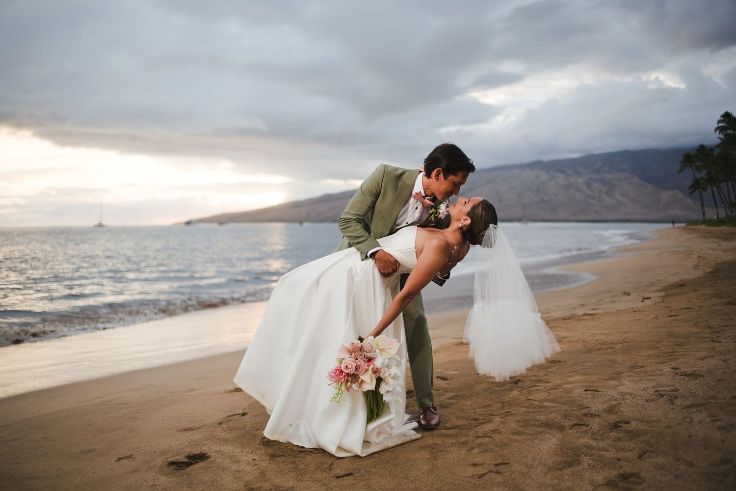 Bride and groom share a romantic dip kiss on a sandy beach at sunset with mountains and ocean in the background.