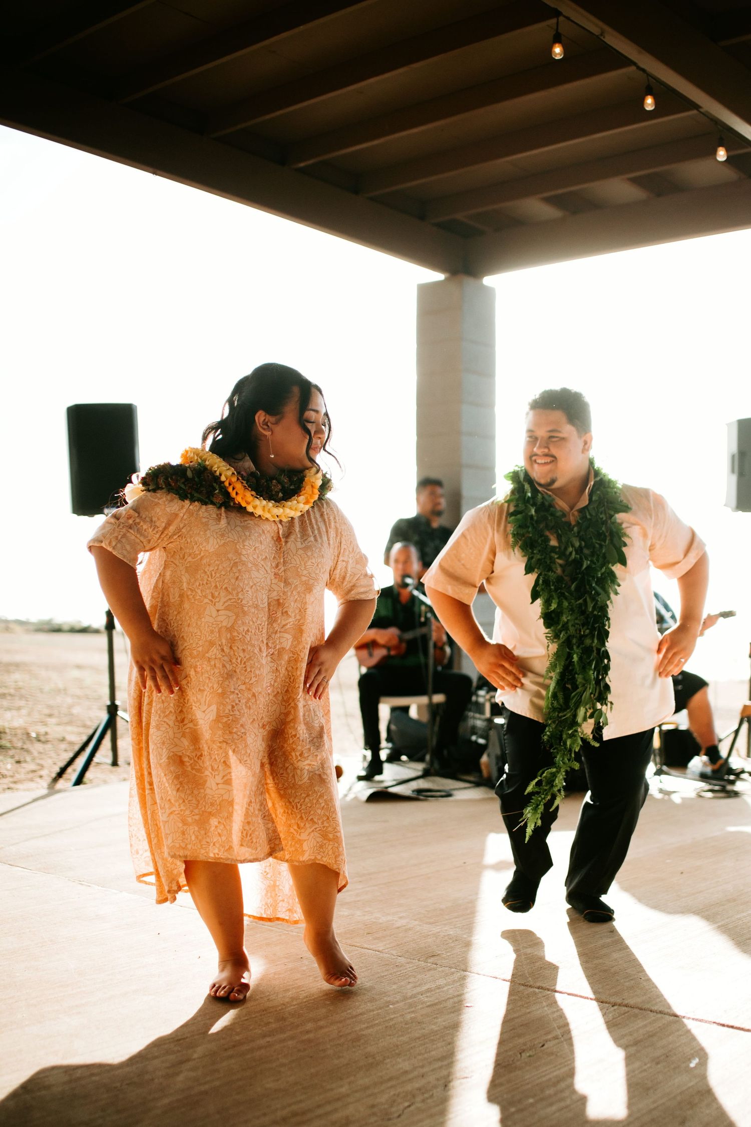 Two hula dancers wearing lei perform at an outdoor Hawaiian celebration with a live band in the background.