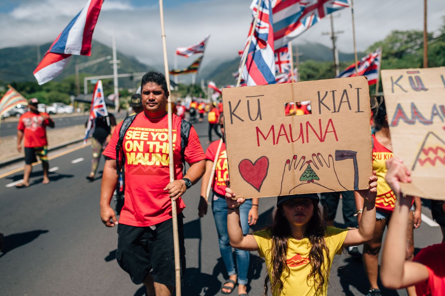 Rally participants hold protest signs and Hawaiian flags while marching on a paved road with mountains behind.