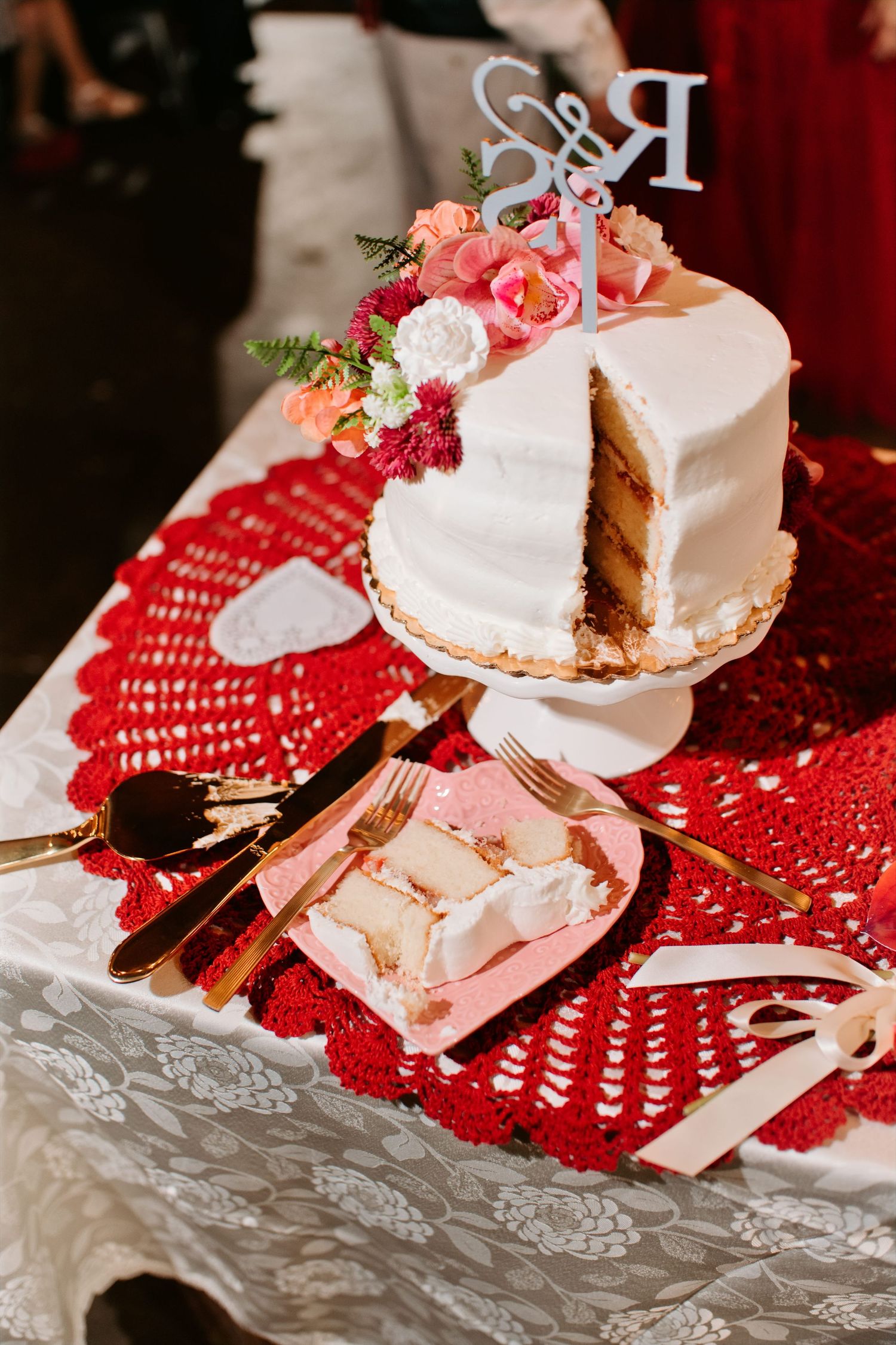White wedding cake with floral topper on red lace tablecloth, with a slice served on a pink plate beside gold cutlery.