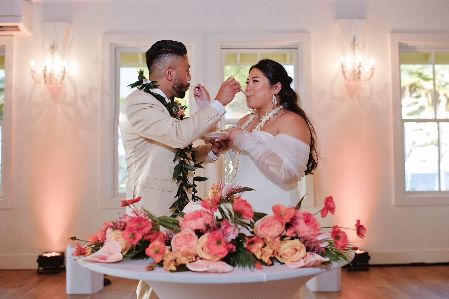 A bride and groom share a joyful cake cutting moment surrounded by pink floral arrangements.