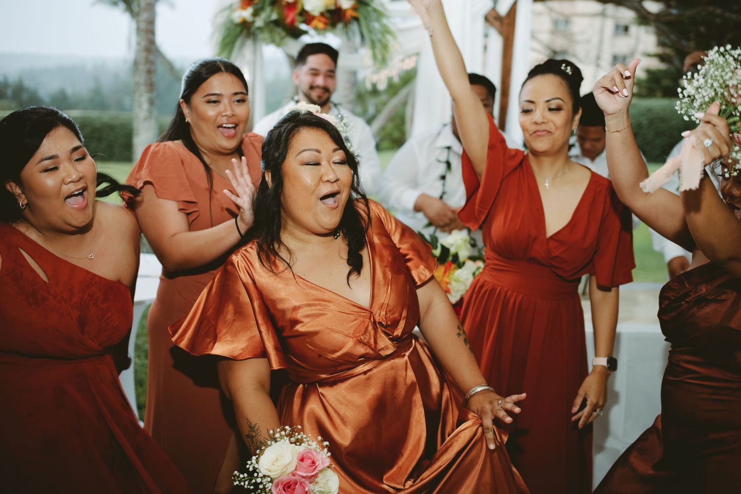 Bridesmaids in rust orange dresses laughing and celebrating joyfully at an outdoor wedding ceremony.