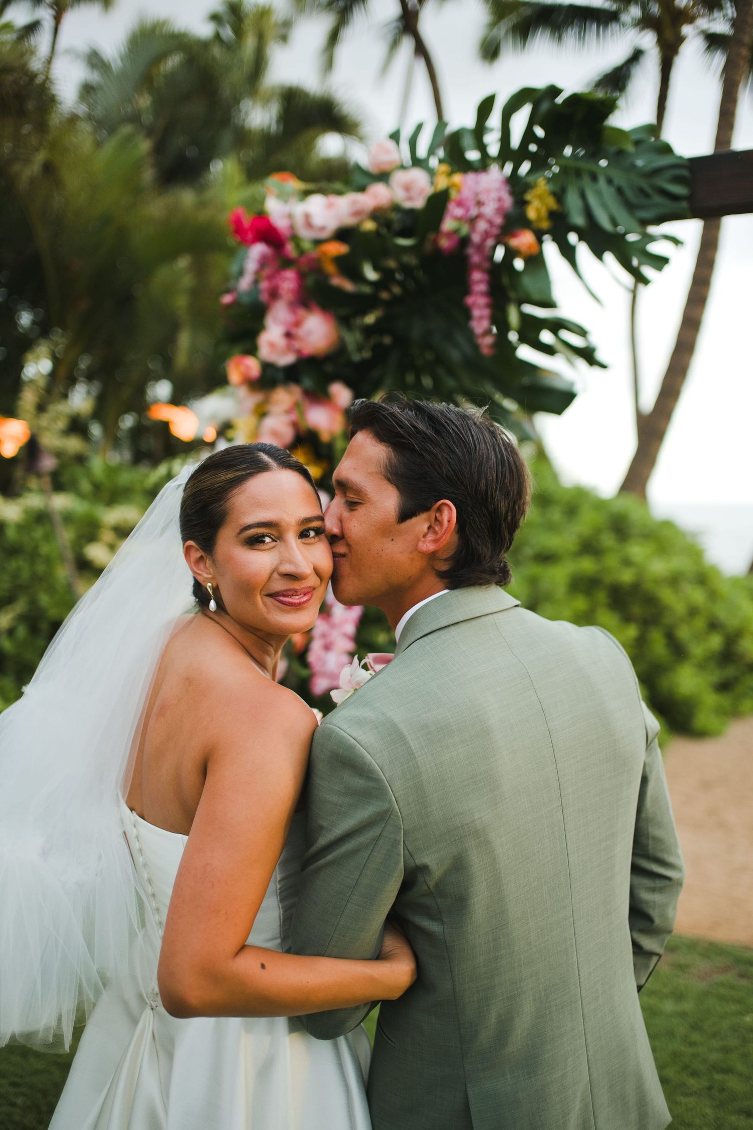 Bride in white gown and veil smiles as groom in sage suit kisses her cheek at tropical outdoor wedding ceremony.