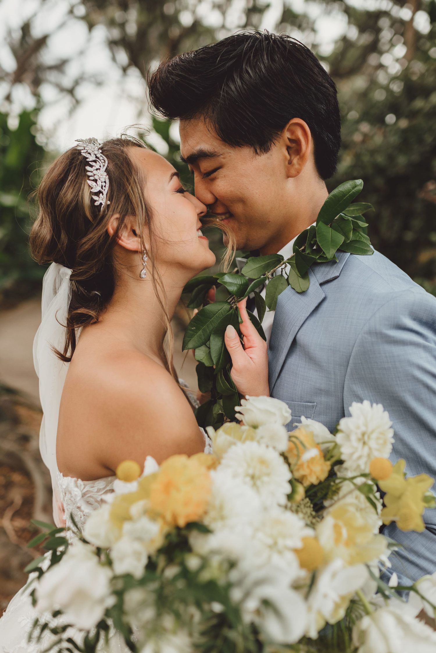 Bride and groom sharing a romantic almost-kiss, she holds white and yellow bouquet, he wears a green lei and gray suit.