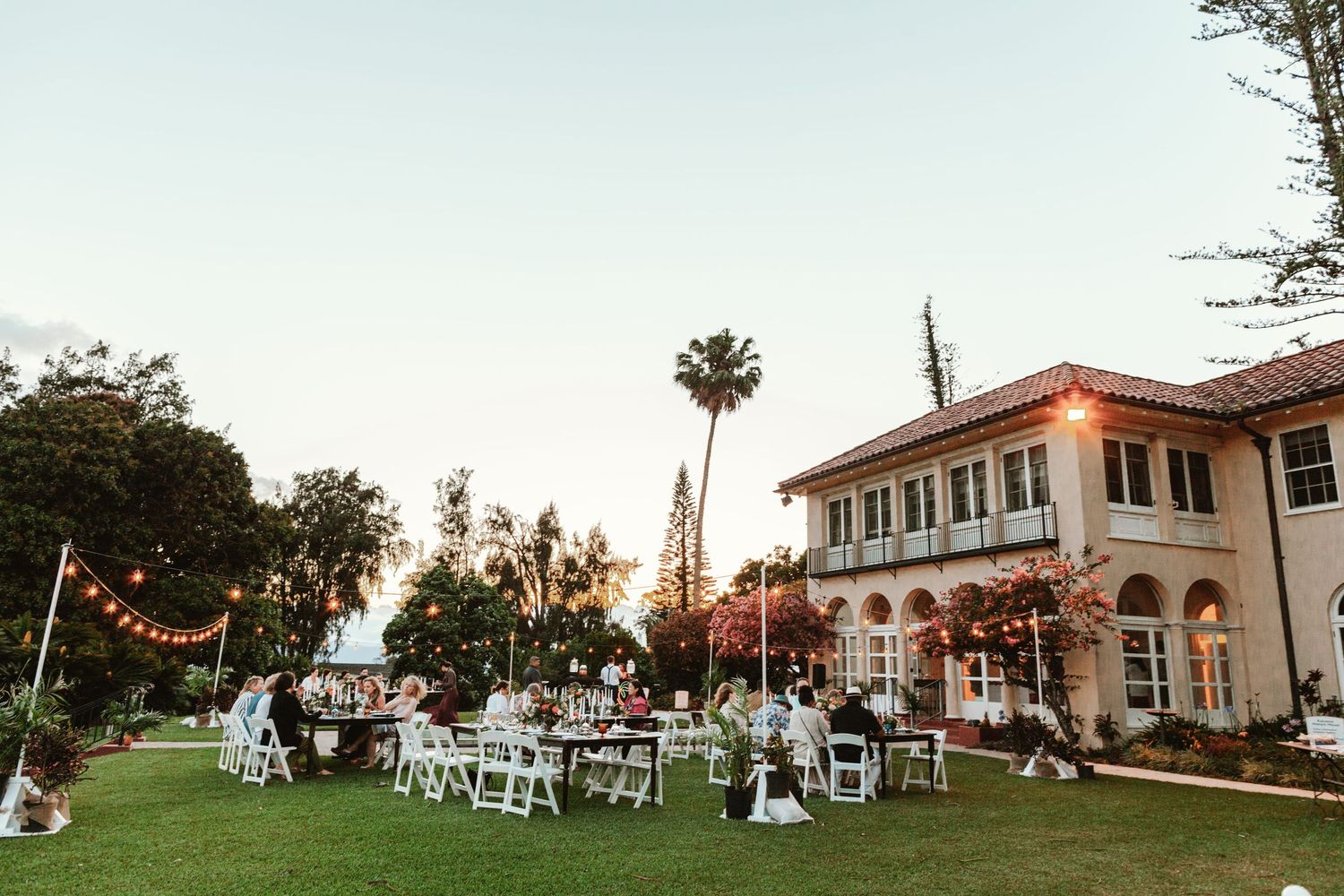 Outdoor wedding reception on lush green lawn at sunset with string lights and elegant white chairs beside a Spanish-style estate.