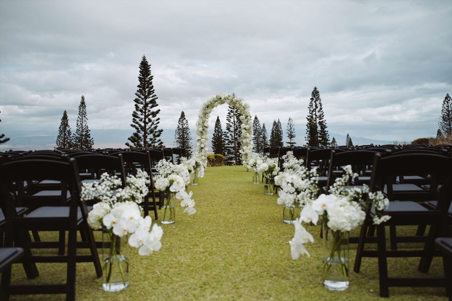Elegant outdoor wedding aisle with white floral arrangements leading to a floral arch amid tall pine trees.