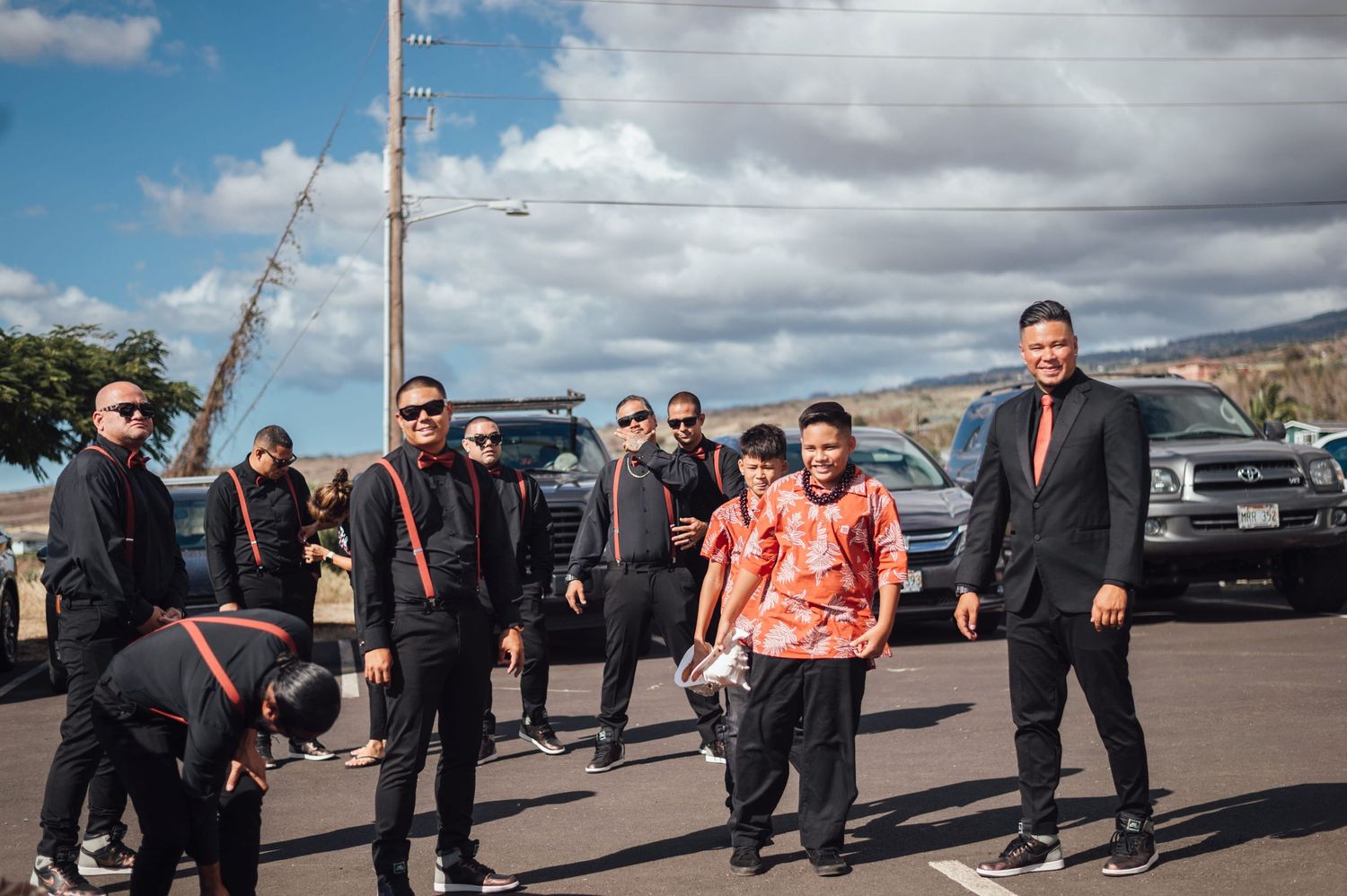 Group of men in black attire posing outdoors in a parking lot on a sunny day with a black SUV in the background.