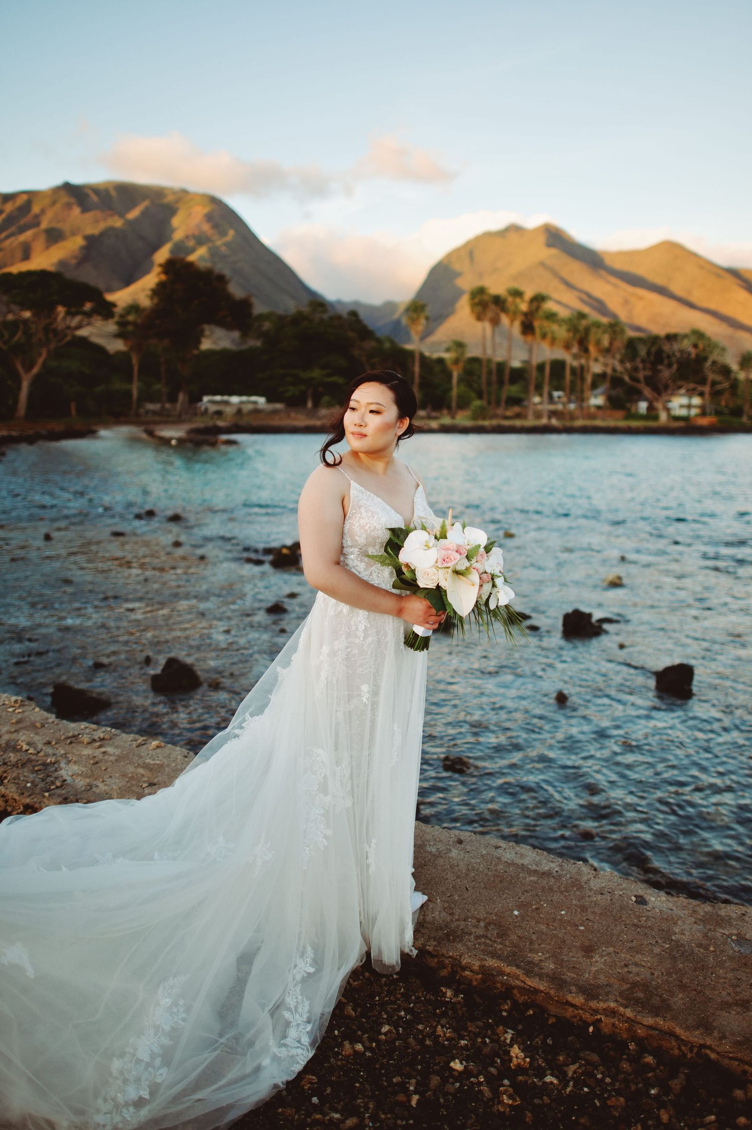 Bride in lace gown holds floral bouquet by Hawaiian coastline with mountains and palm trees at golden hour.
