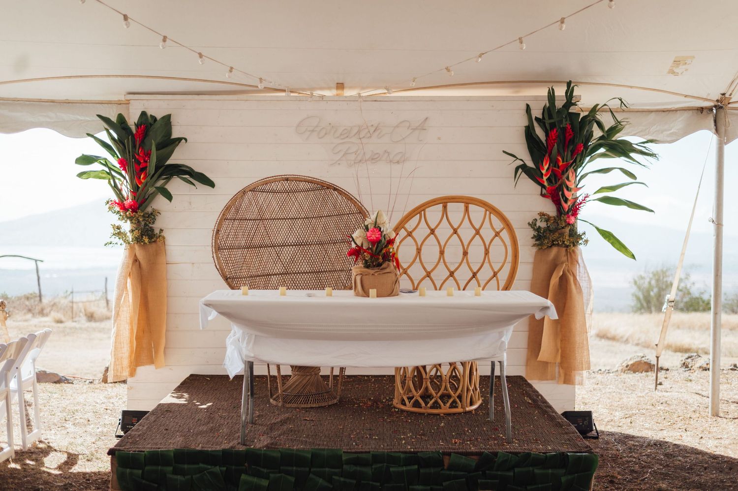 Boho desert wedding sweetheart table with rattan chairs, tropical florals, and a white shiplap backdrop under a tent.