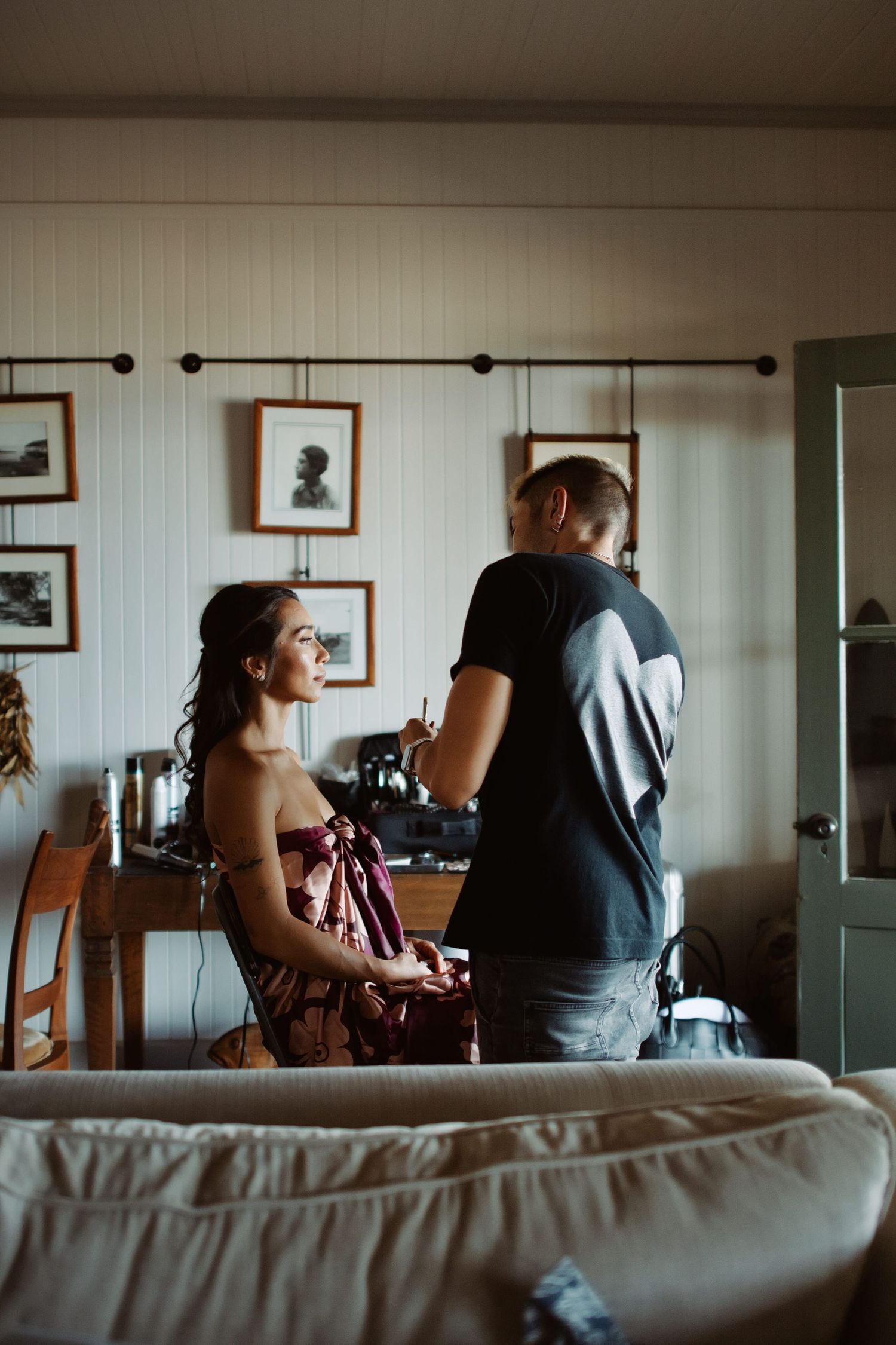 A woman in a strapless dress sits while a man in a black t-shirt attends to her hair in a cozy room.