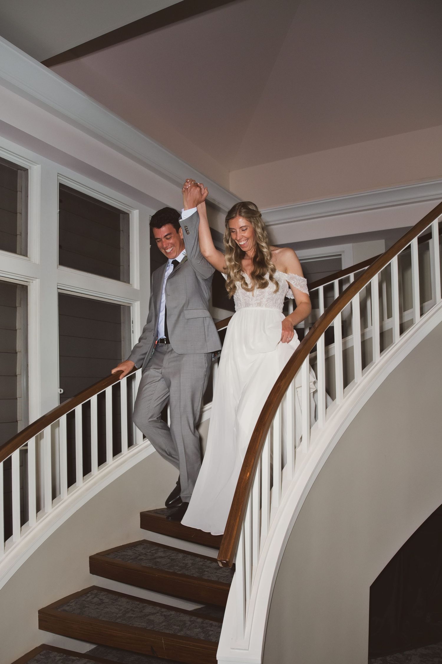 Bride and groom holding hands walking down elegant curved staircase, wearing white gown and gray suit.