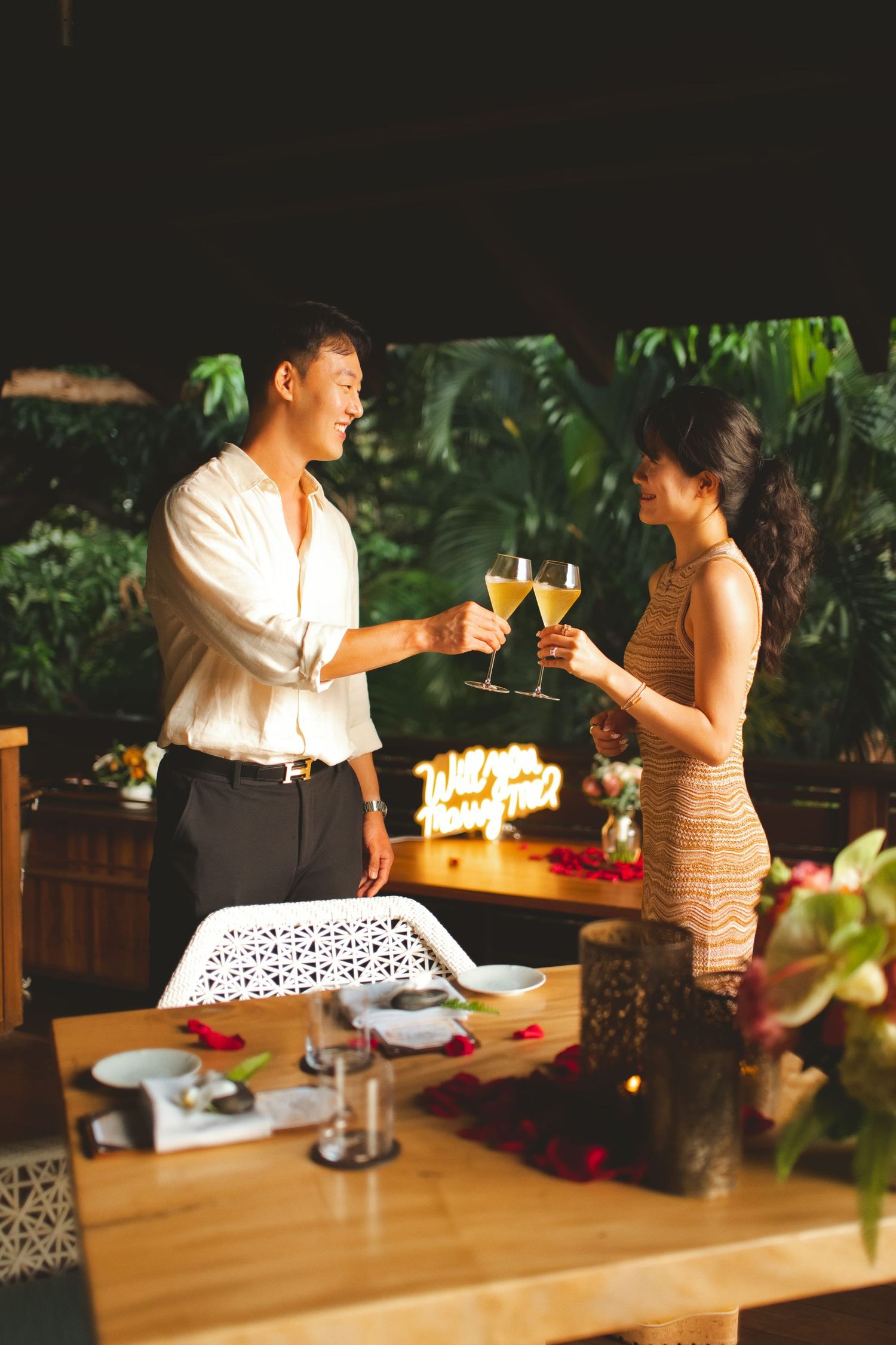 Couple toasting with champagne glasses at a romantic candlelit dinner surrounded by tropical greenery.