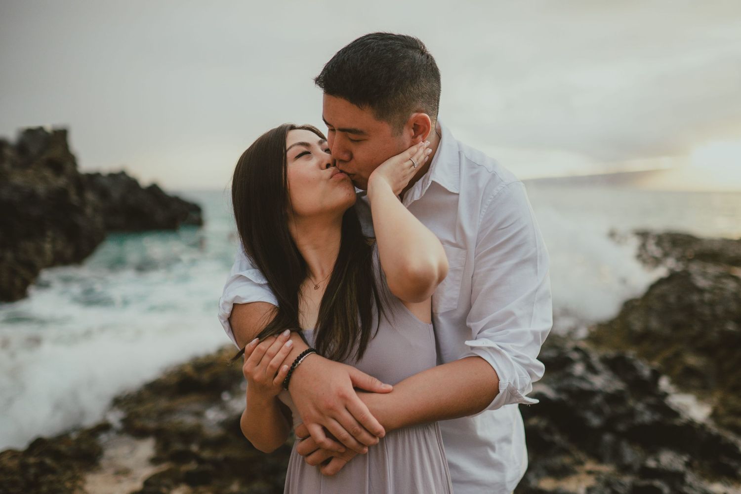 Couple sharing romantic kiss on rocky Hawaiian coastline with crashing ocean waves.