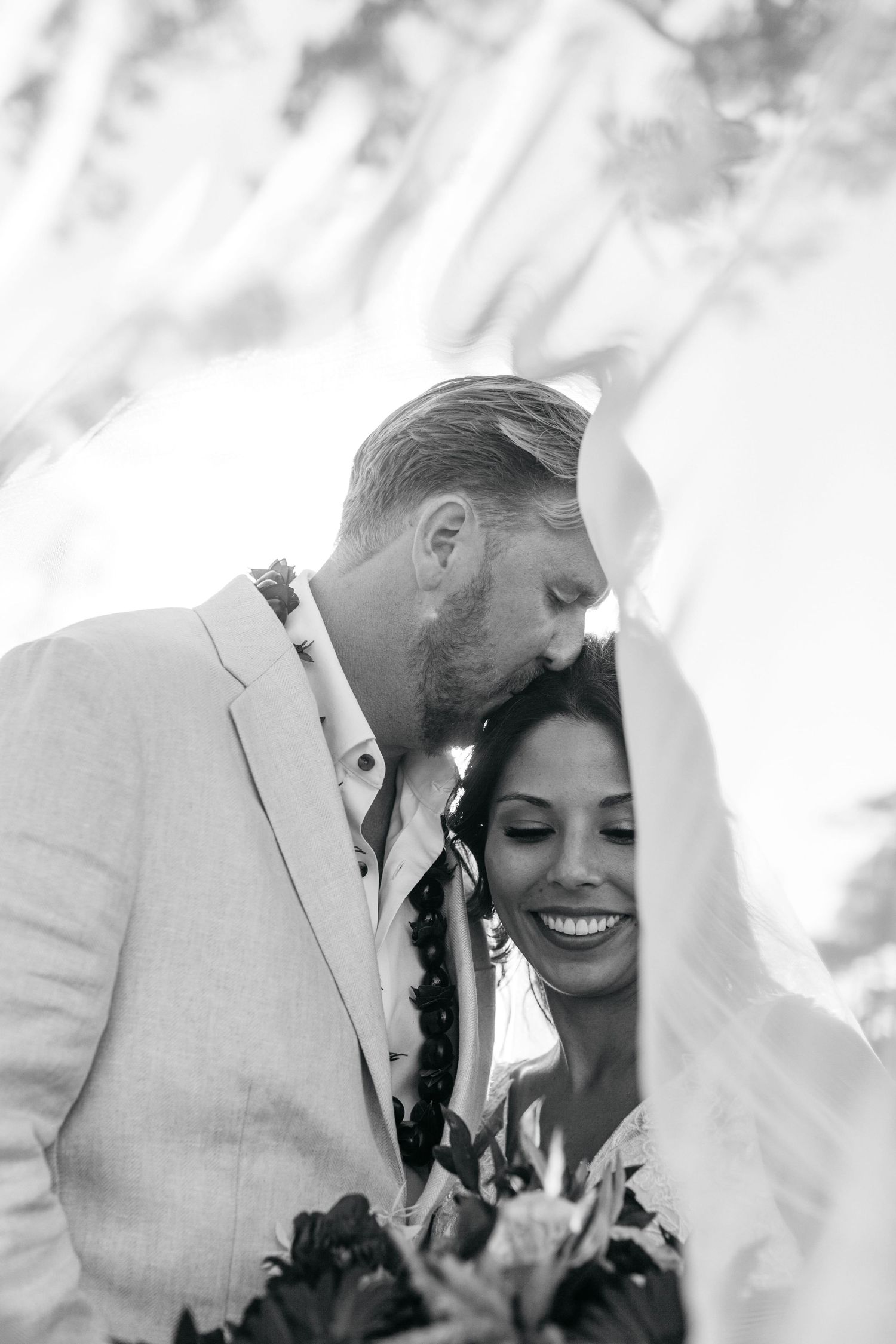 Black and white photo of groom kissing bride on head, both wearing leis, bride smiling holding tropical bouquet.