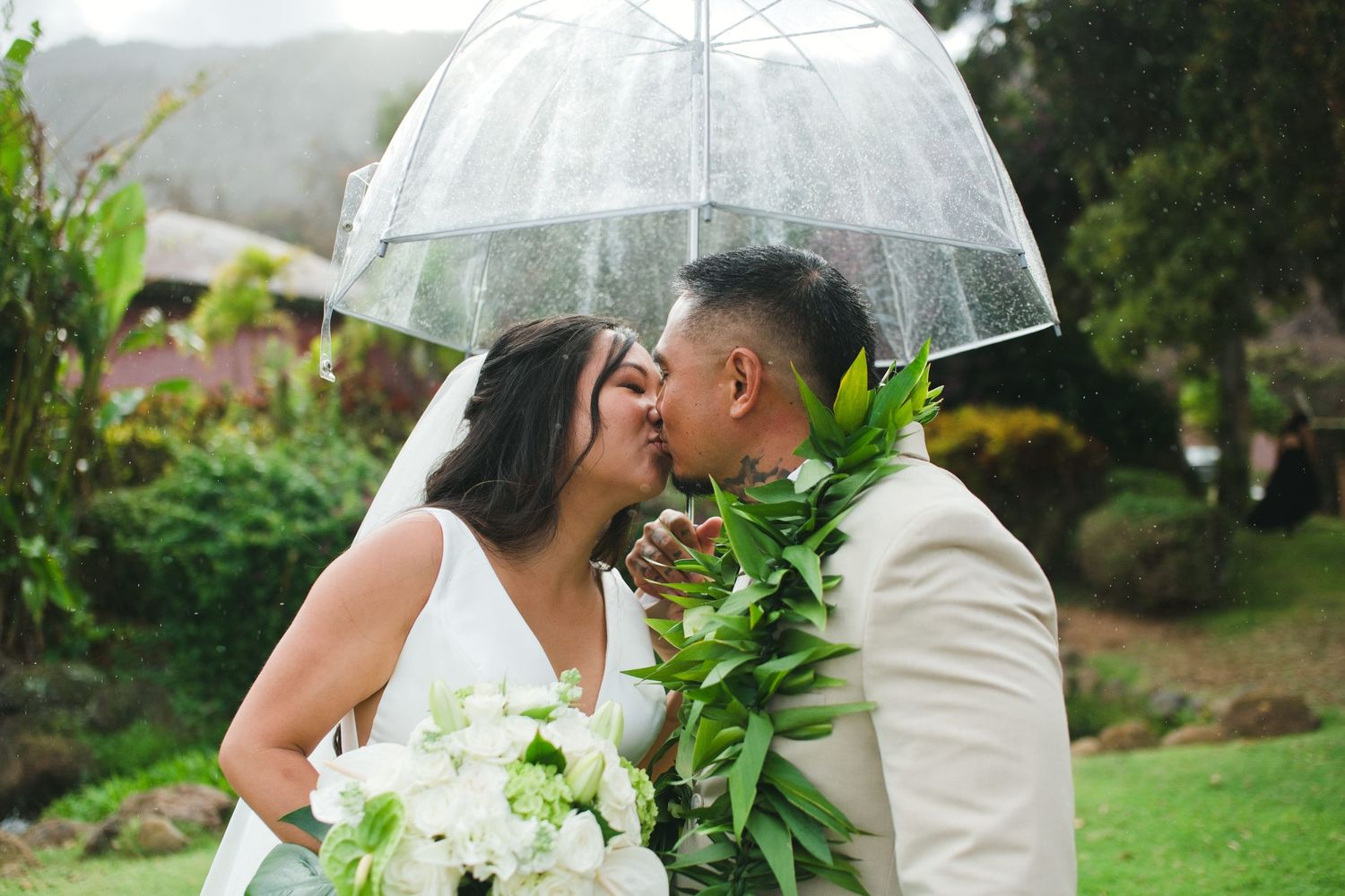Bride and groom kissing under a clear umbrella in the rain, bride holding white bouquet, groom wearing green lei.