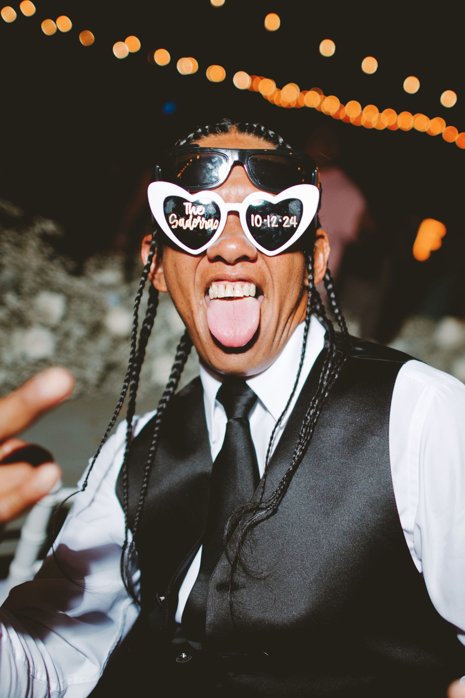 Person in heart-shaped wedding sunglasses dated 10-12-24 with braids, sticking tongue out at a wedding reception.