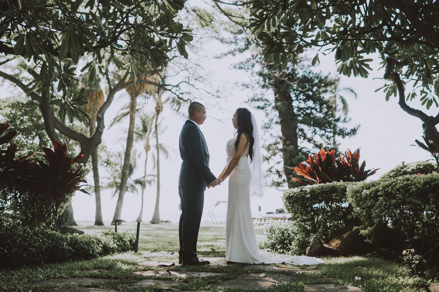 Bride and groom holding hands in a lush tropical garden ceremony surrounded by palm trees and vibrant greenery.