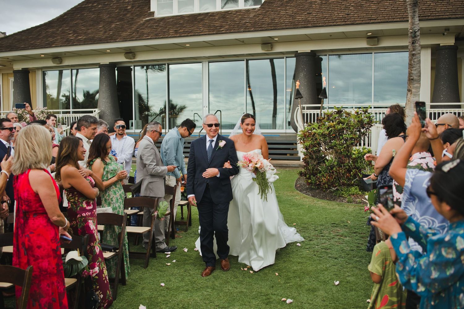 Bride walks down outdoor aisle with escort at tropical wedding venue, guests celebrating with flower petals.