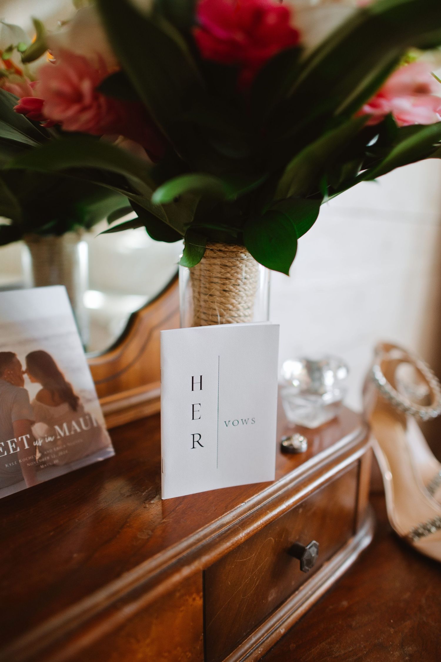 Wedding vows booklet labeled HER displayed on wooden dresser beside bridal bouquet and heels.