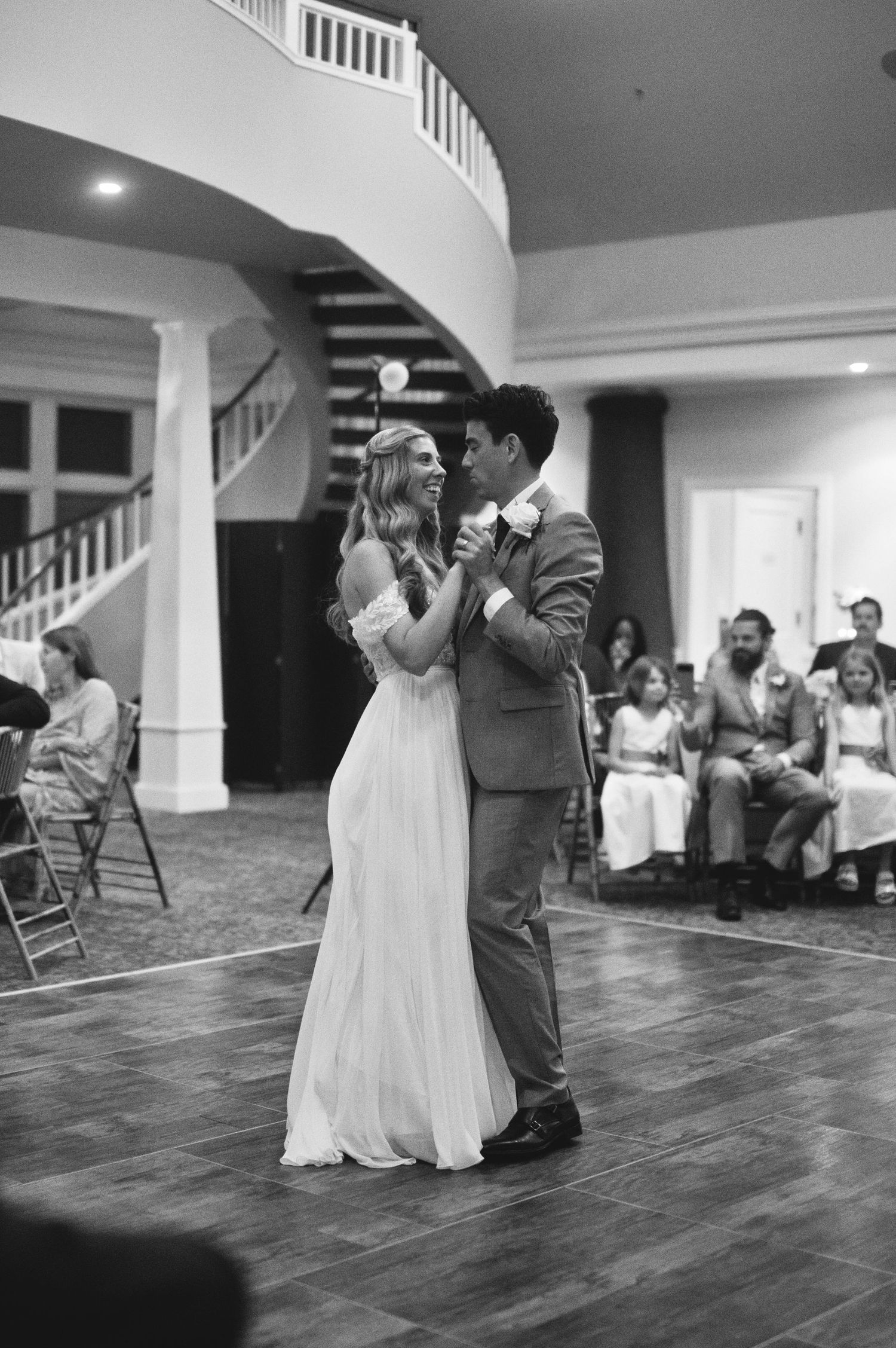 Bride and groom share their first dance at wedding reception in elegant ballroom with grand staircase.