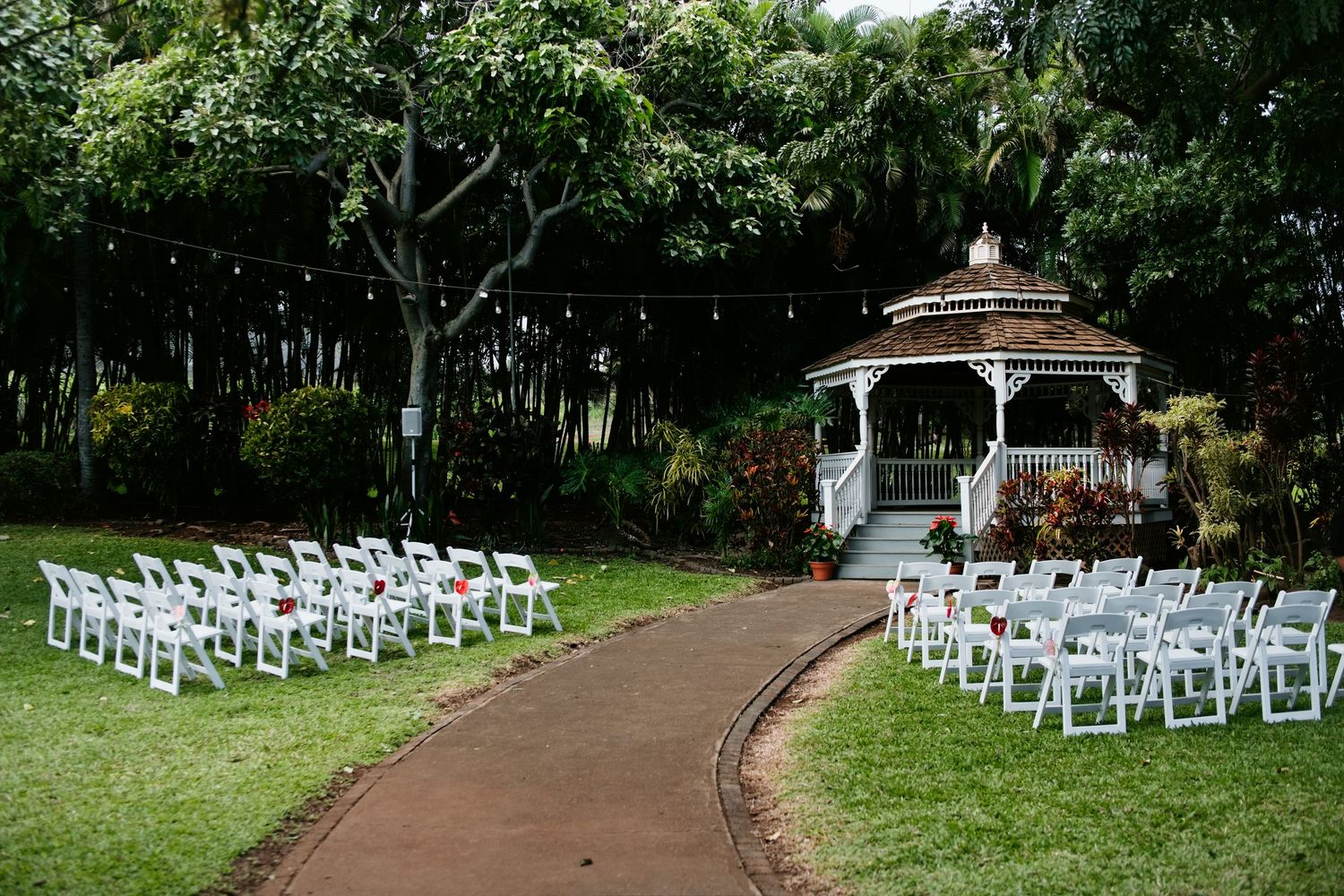 Outdoor wedding ceremony setup with white chairs lining a path leading to a gazebo surrounded by lush tropical greenery.