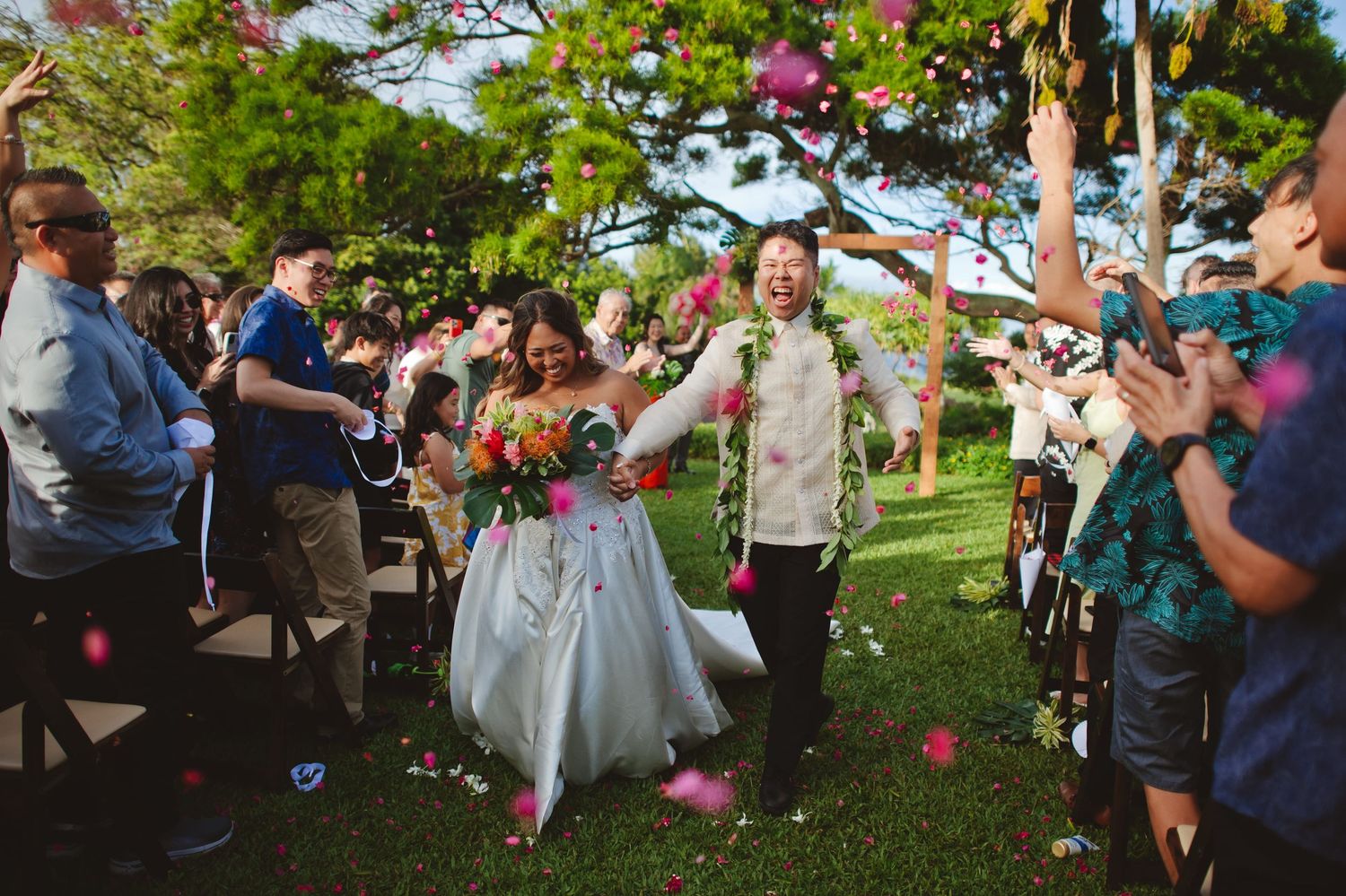 Joyful newlyweds walk down outdoor aisle as guests toss colorful flower petals at garden wedding ceremony.