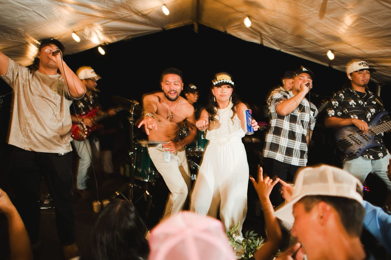 Bride and groom dance joyfully with guests at lively outdoor Hawaiian wedding reception.