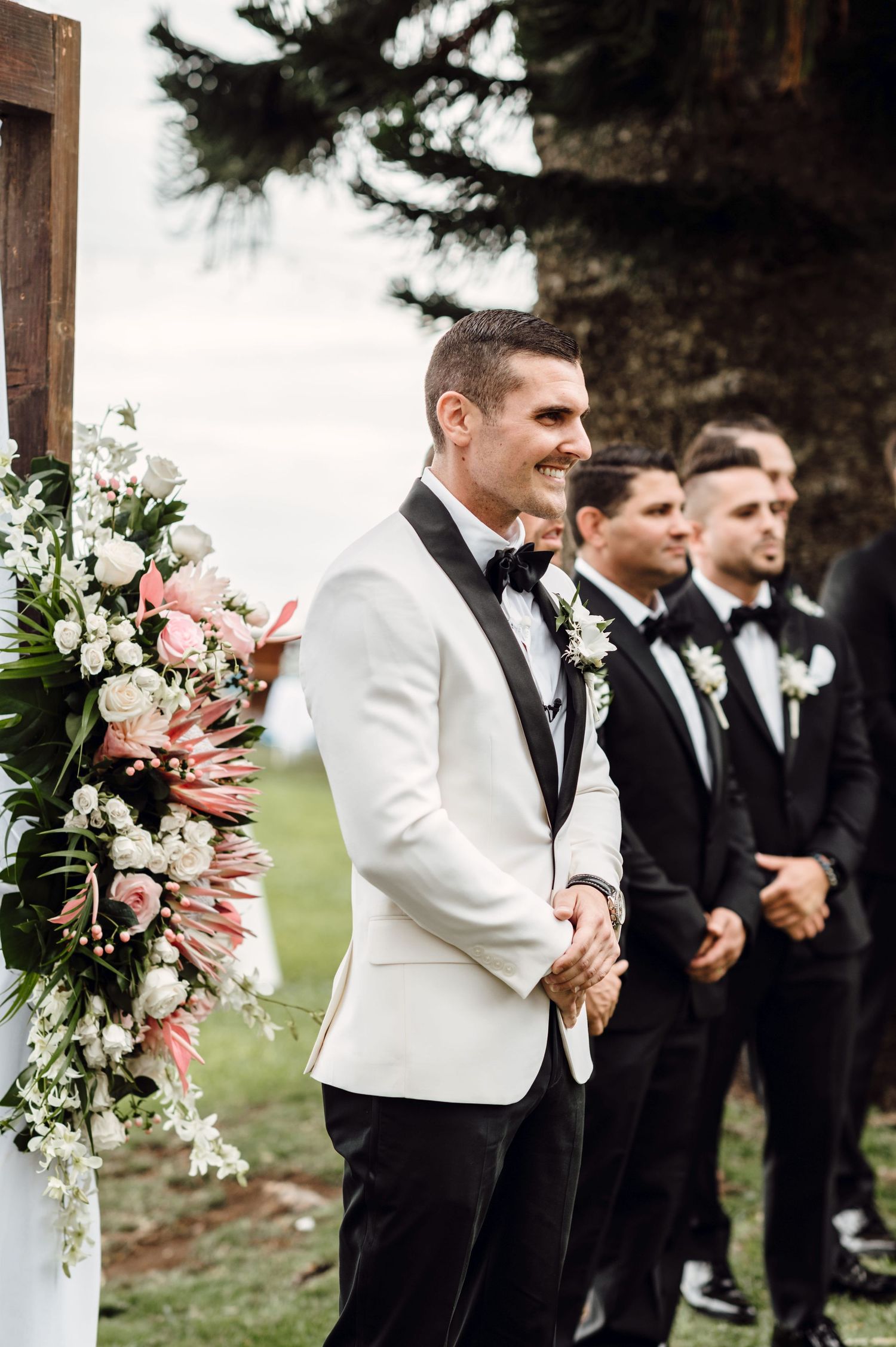 Smiling groom in white tuxedo jacket stands at wedding altar with groomsmen in black suits and floral arrangements.