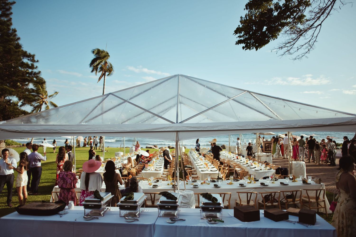 Outdoor beachside wedding reception under a clear tent with long dining tables and tropical palm trees.