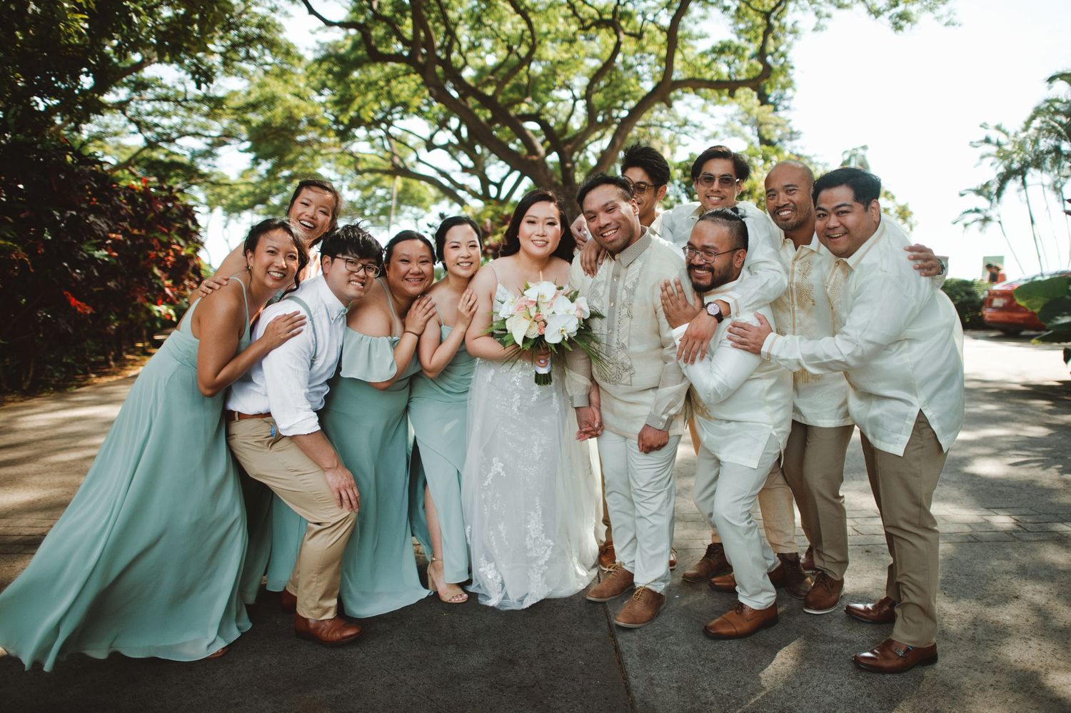 Wedding party posing joyfully outdoors, bride in white gown with bridesmaids in sage green dresses and groomsmen in white shirts.