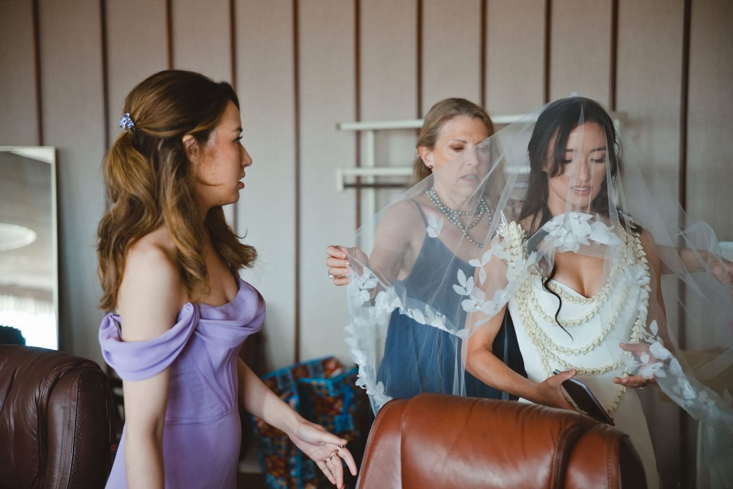 Bridesmaids in purple and blue dresses help a bride with her wedding veil during bridal preparations.