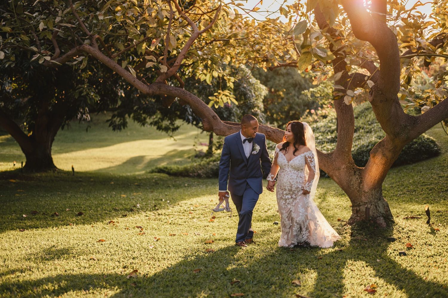 Bride and groom walking hand in hand under a sunlit tree on lush green grass during golden hour.