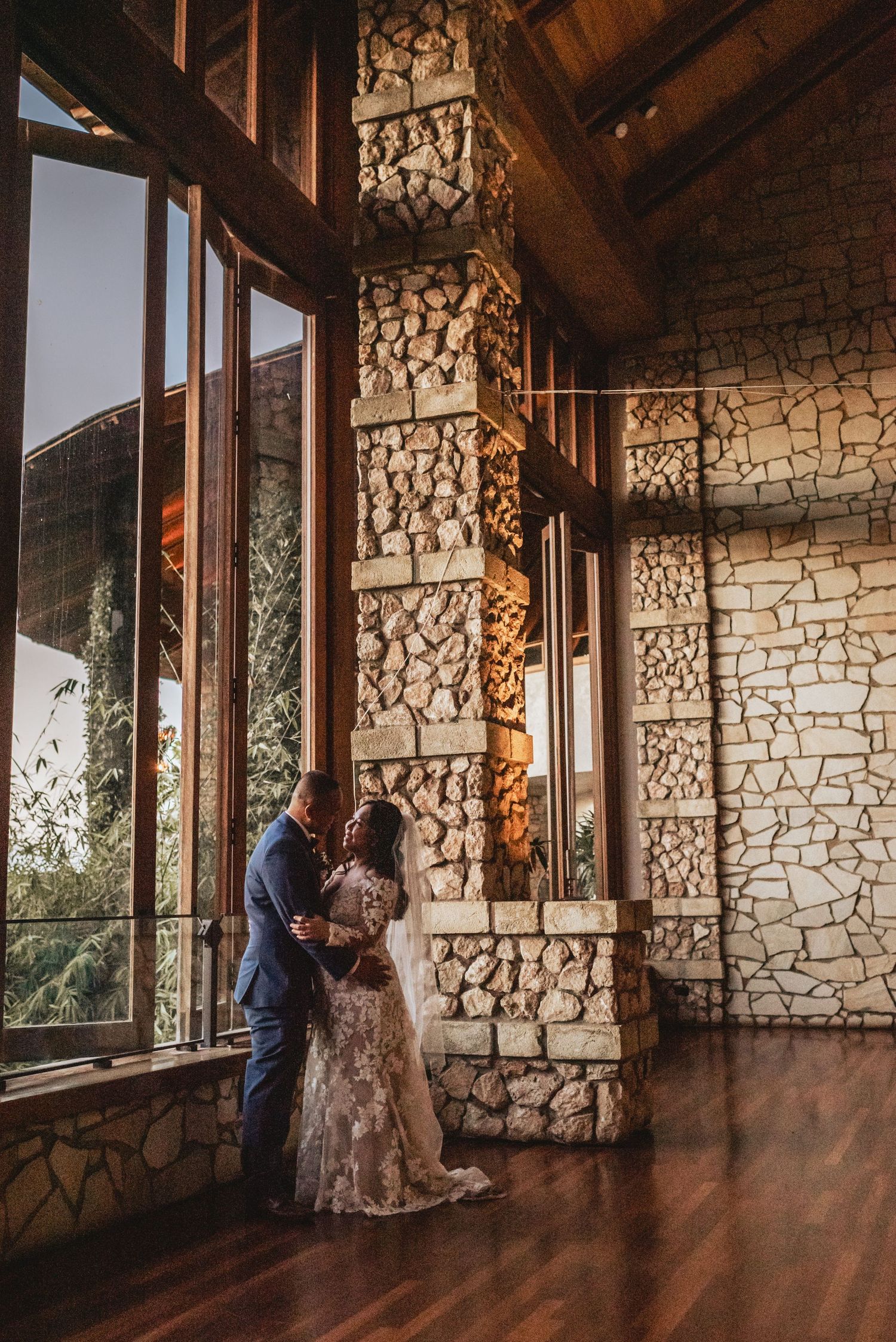 Bride and groom share a romantic embrace beside stone pillars in an elegant rustic wedding venue.
