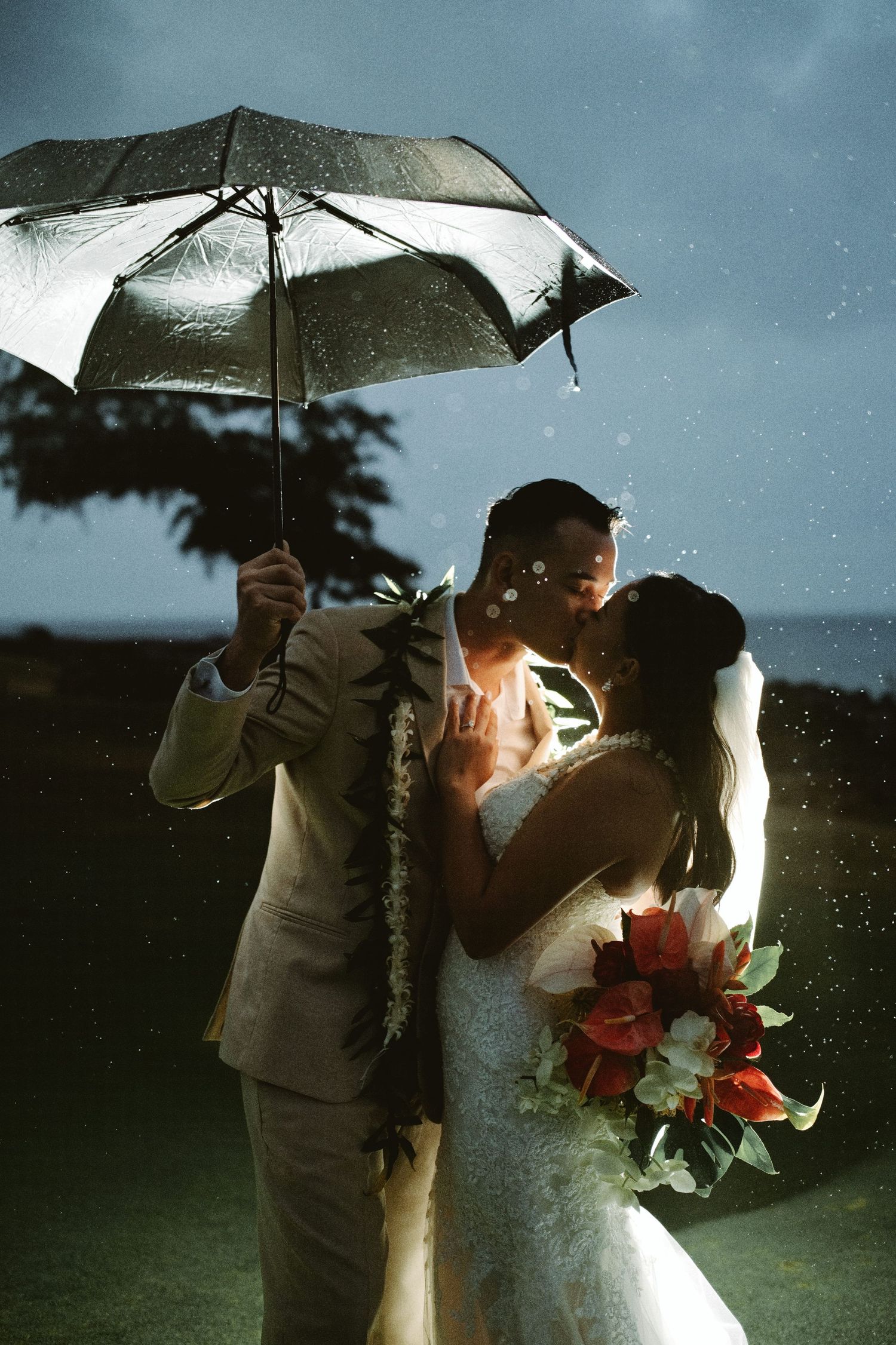 Bride and groom kissing under umbrella in rain, bride holding red bouquet, romantic wedding portrait.