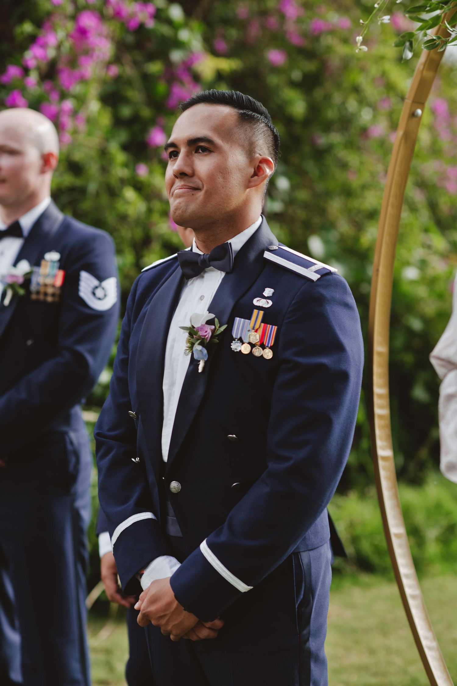 Military groom in dress uniform with medals smiles emotionally at outdoor wedding ceremony.