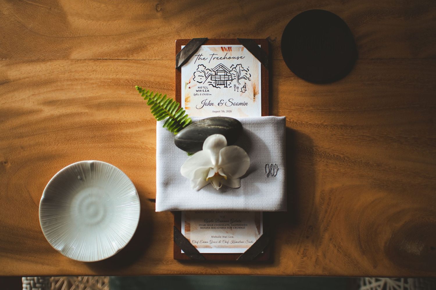 Elegant wedding place setting with white napkin, garlic flower, fern, and calligraphy menu card on wooden table.