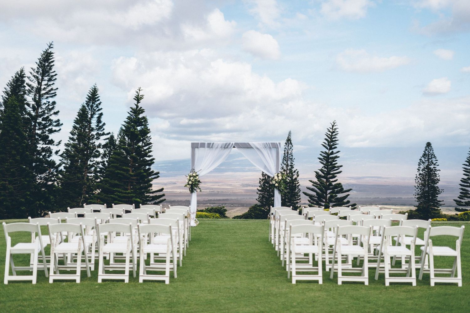 Outdoor wedding ceremony setup with white chairs and arch overlooking mountain landscape.