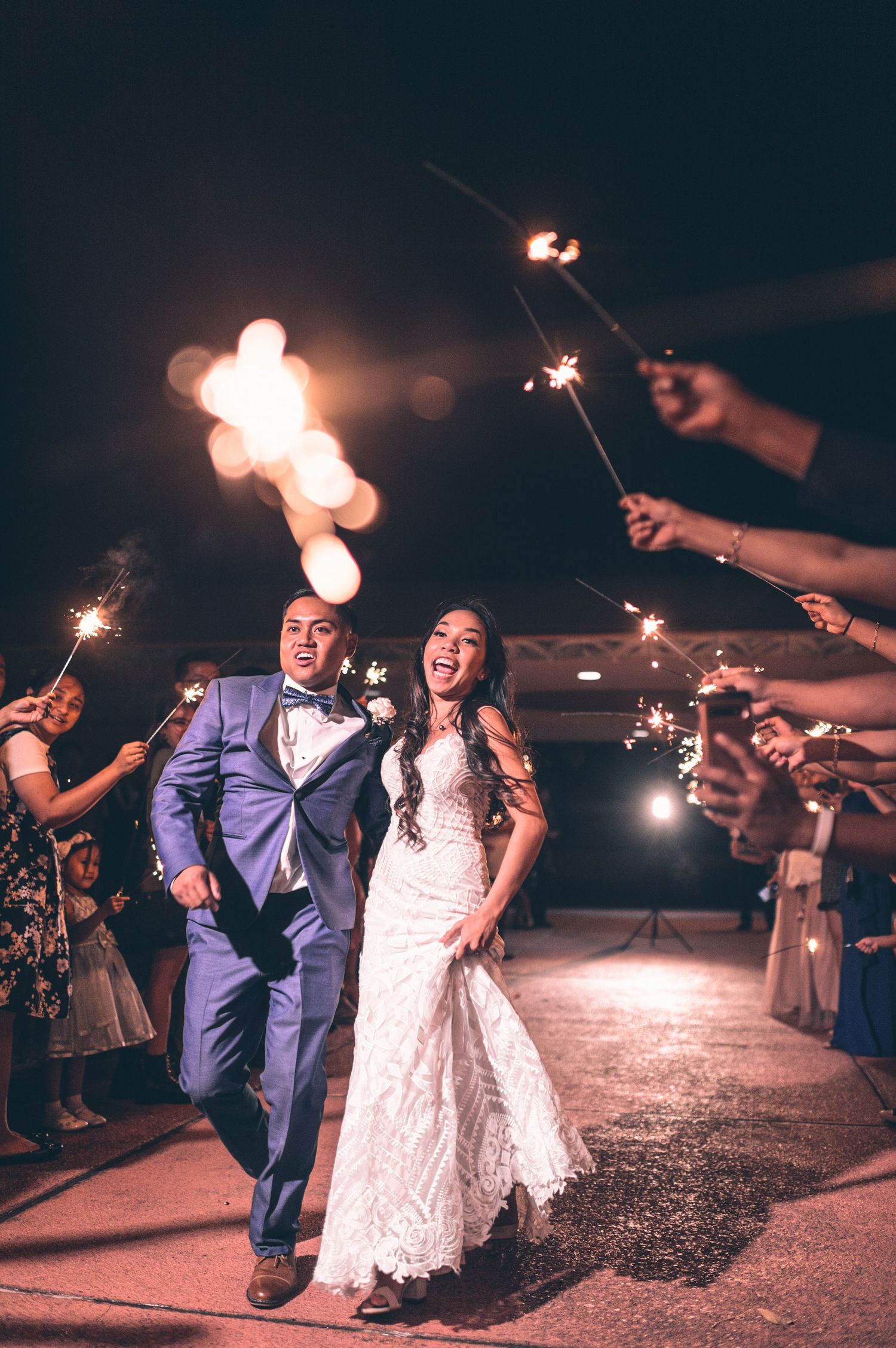 Wedding couple makes grand exit through sparkler tunnel held by guests at night.