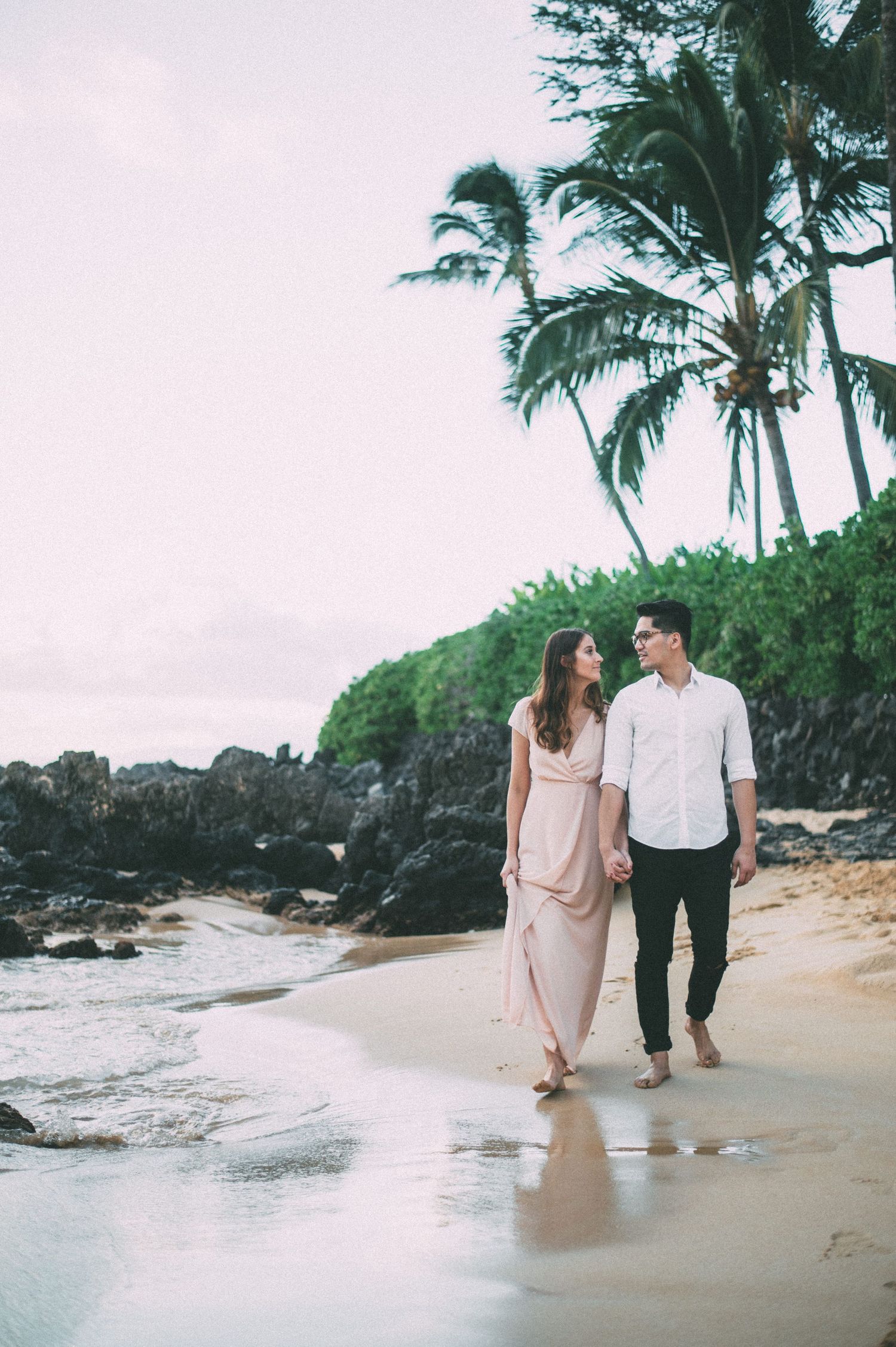 Couple holding hands and walking along a tropical beach with palm trees at dusk.