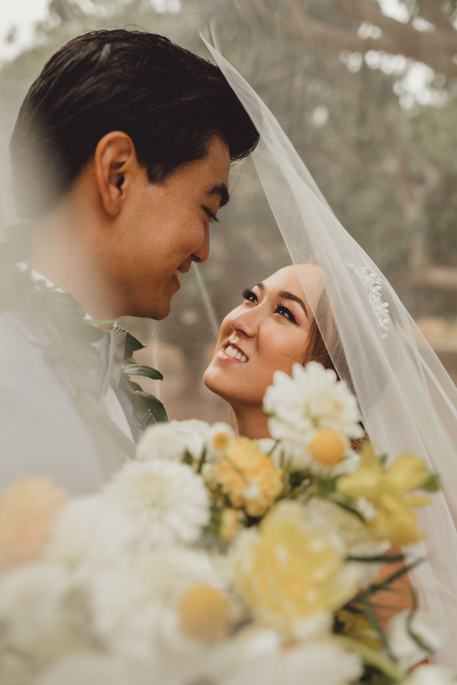 A smiling bride and groom share an intimate moment under a wedding veil, with a bouquet of white and yellow flowers.