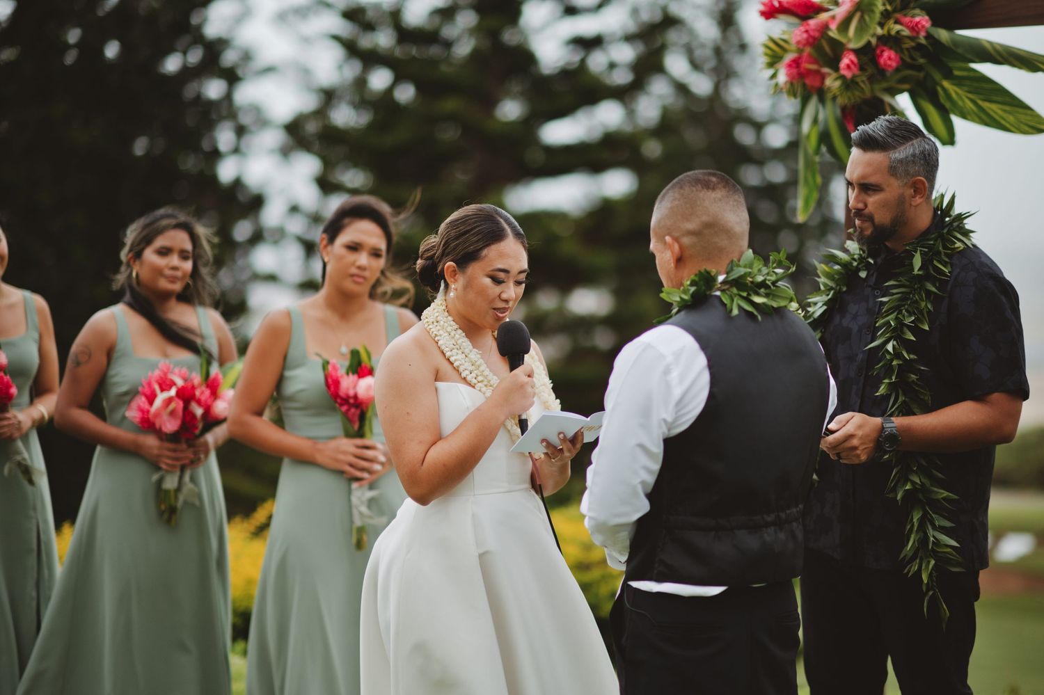 Bride and groom exchange vows outdoors with bridesmaids in sage green dresses holding red bouquets at Hawaiian wedding.