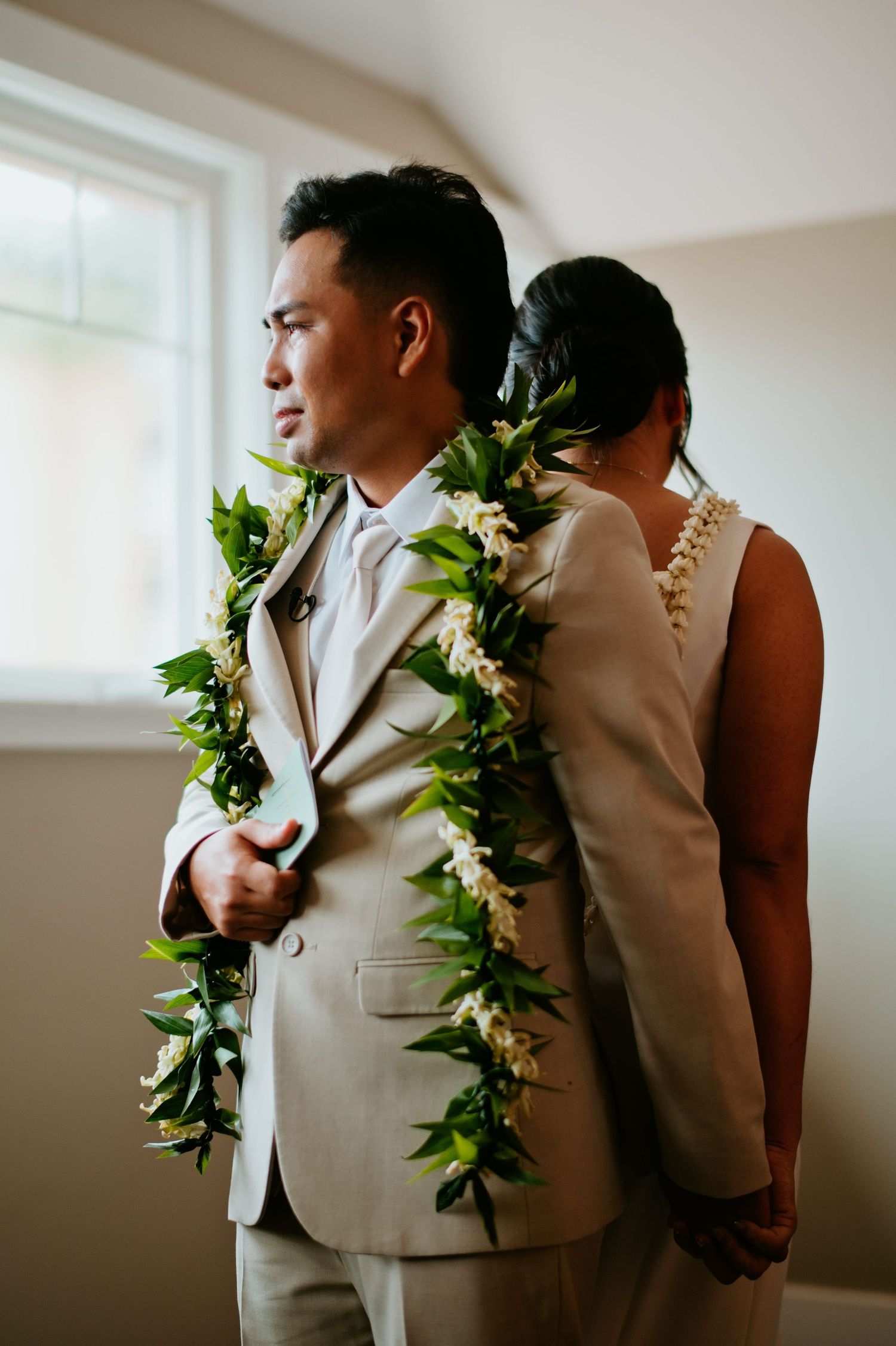 Couple in floral leis sharing a tender back-to-back moment, groom in tan suit near window.