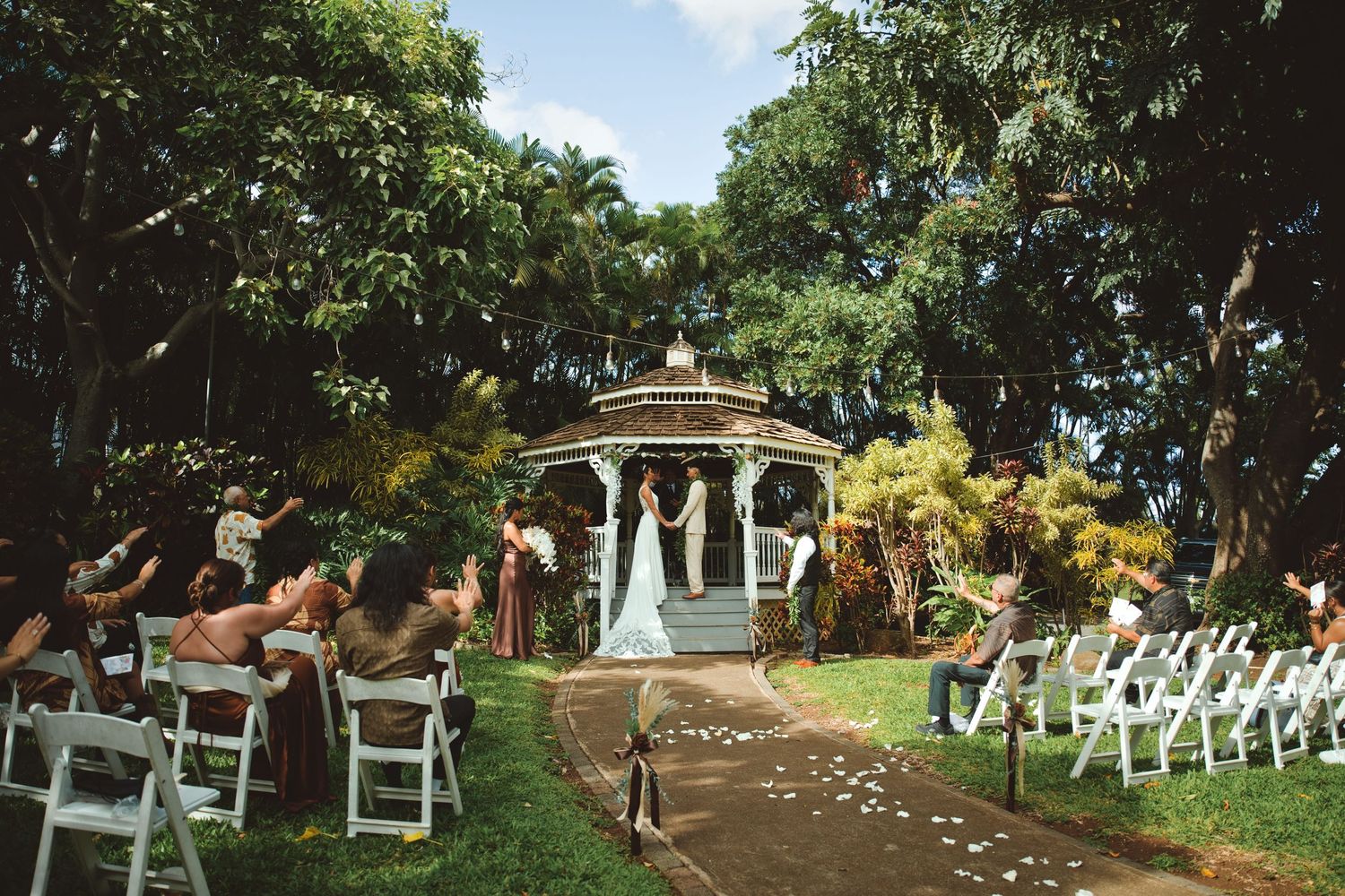 Outdoor wedding ceremony at a gazebo surrounded by lush tropical trees with white chairs and rose petal aisle.