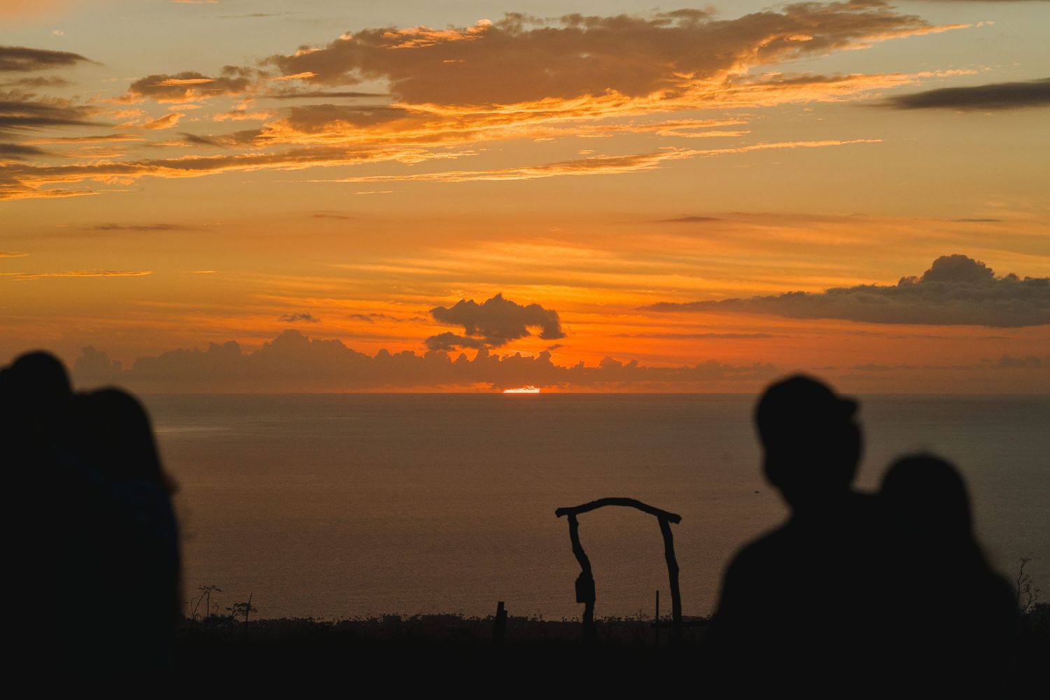 Silhouettes of people watching a vibrant orange sunset over the ocean with dramatic clouds in the sky.