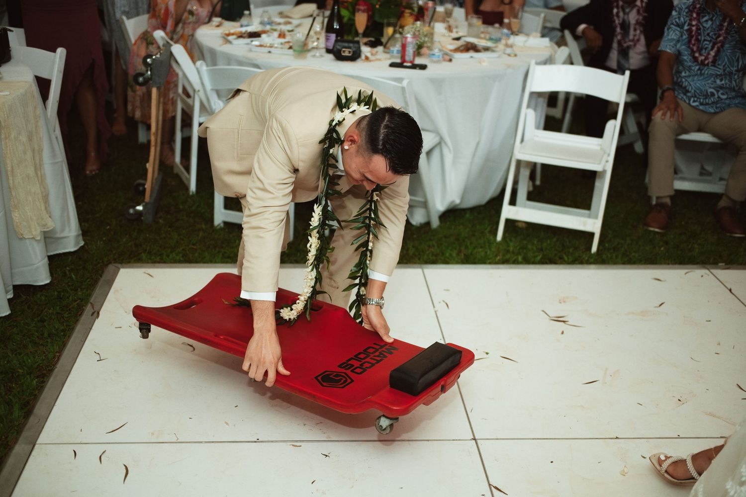 Man in suit with lei bends over red mechanic creeper on white dance floor at outdoor wedding reception.