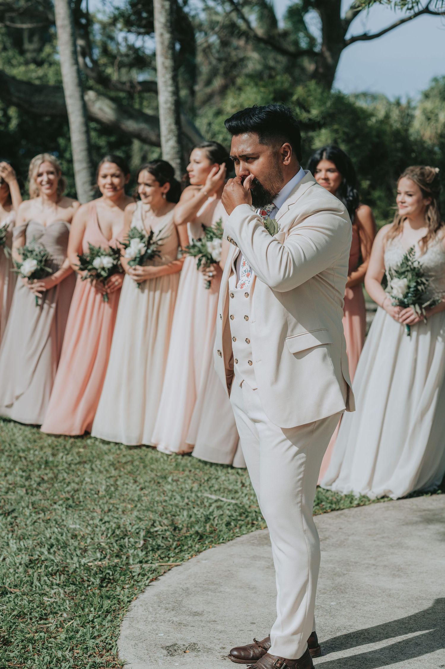 Emotional groom in white suit covering his mouth as bridesmaids in blush pink dresses line up behind him at outdoor wedding.